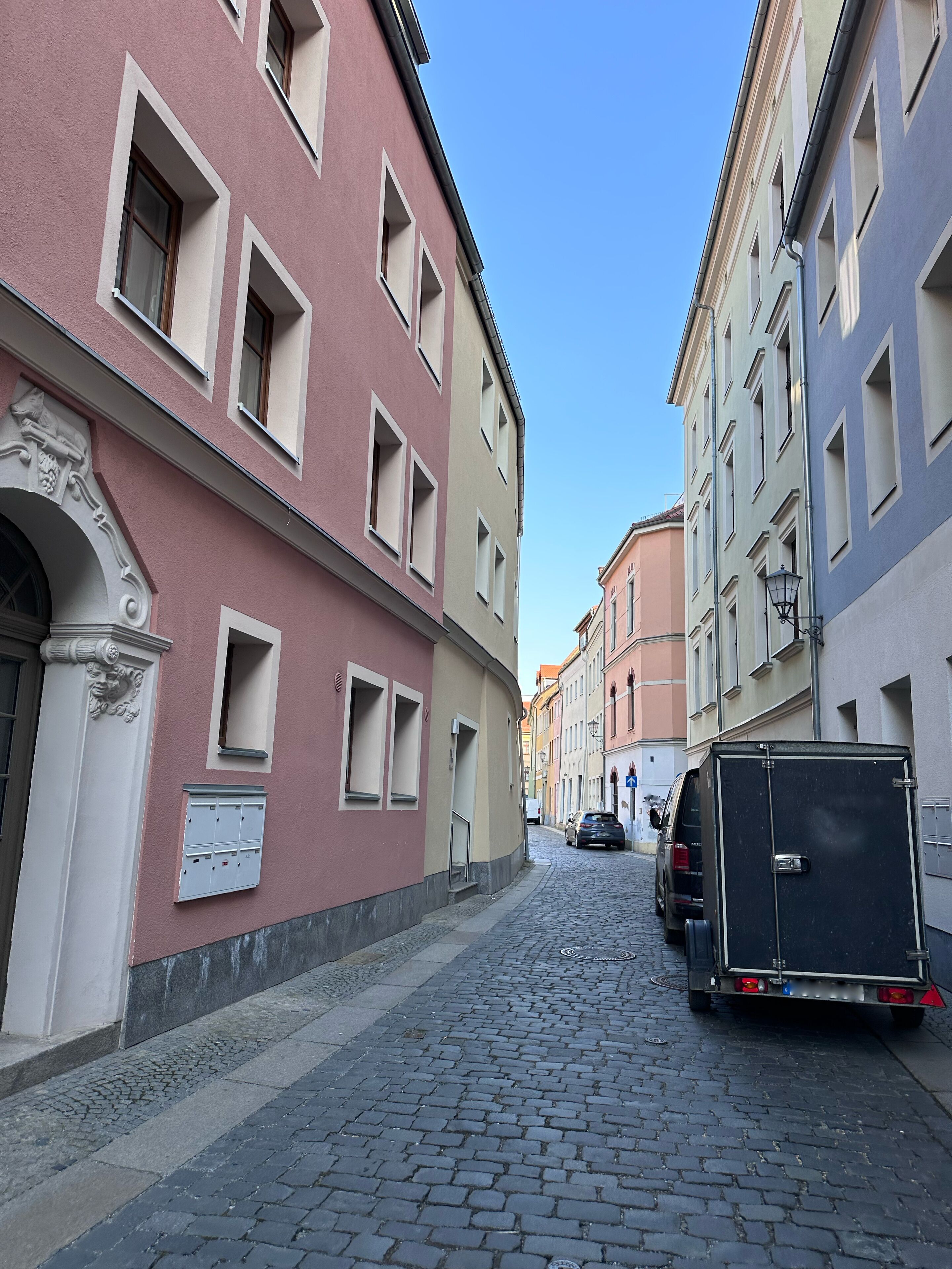 A narrow cobblestone street flanked by historic buildings with uniform window designs and a black delivery truck parked on the right.