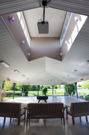 An indoor patio with a high ceiling, recessed lighting, and a projector. There are wooden benches and a grill area.