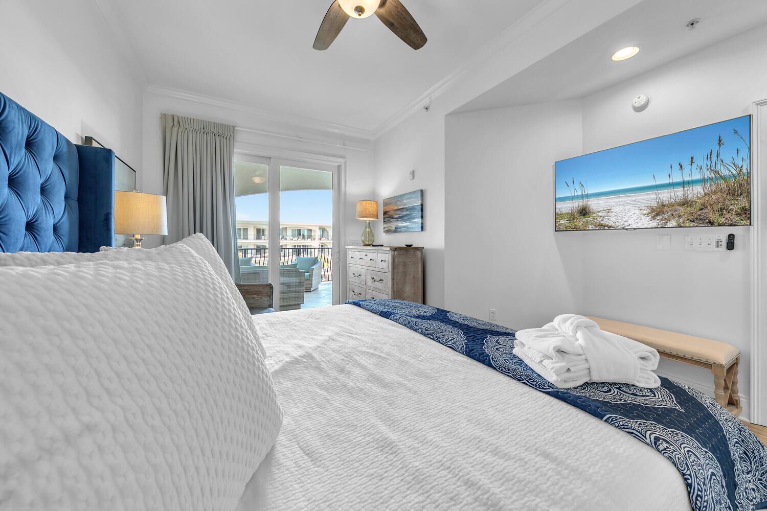 A hotel room with a large bed, a ceiling fan, a TV, and a painting of a beach scene.
