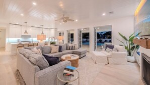 A spacious living area with a large gray sectional sofa, a round coffee table, and a fireplace. There is a kitchen in the background with white cabinets and pendant lighting. - Steps to Vanderbilt Beach, Sleeps 13, Resort-Style Backyard (Naples)