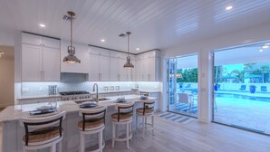A modern kitchen with white cabinetry, a central island with bar stools, and a view of a swimming pool. - Steps to Vanderbilt Beach, Sleeps 13, Resort-Style Backyard (Naples)
