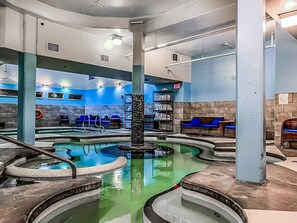 Une piscine intérieure dotée de plusieurs couloirs, d’une fontaine centrale et d’espaces de détente avec des coussins bleus.