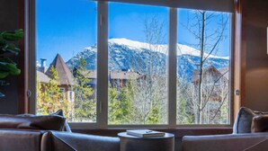 A room with a large window offering a mountain view, a sofa, and a small table with a book. - Mountain View Stoneridge - Pool & Hot Tub & Sauna (Canmore)