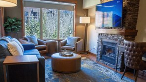 A cozy living room with a fireplace, leather sofas, a coffee table, and a large window offering a view of trees. - Mountain View Stoneridge - Pool & Hot Tub & Sauna (Canmore)