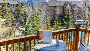 A wooden deck with a table and chairs, overlooking a mountainous landscape and a row of buildings. - Mountain View Stoneridge - Pool & Hot Tub & Sauna (Canmore)