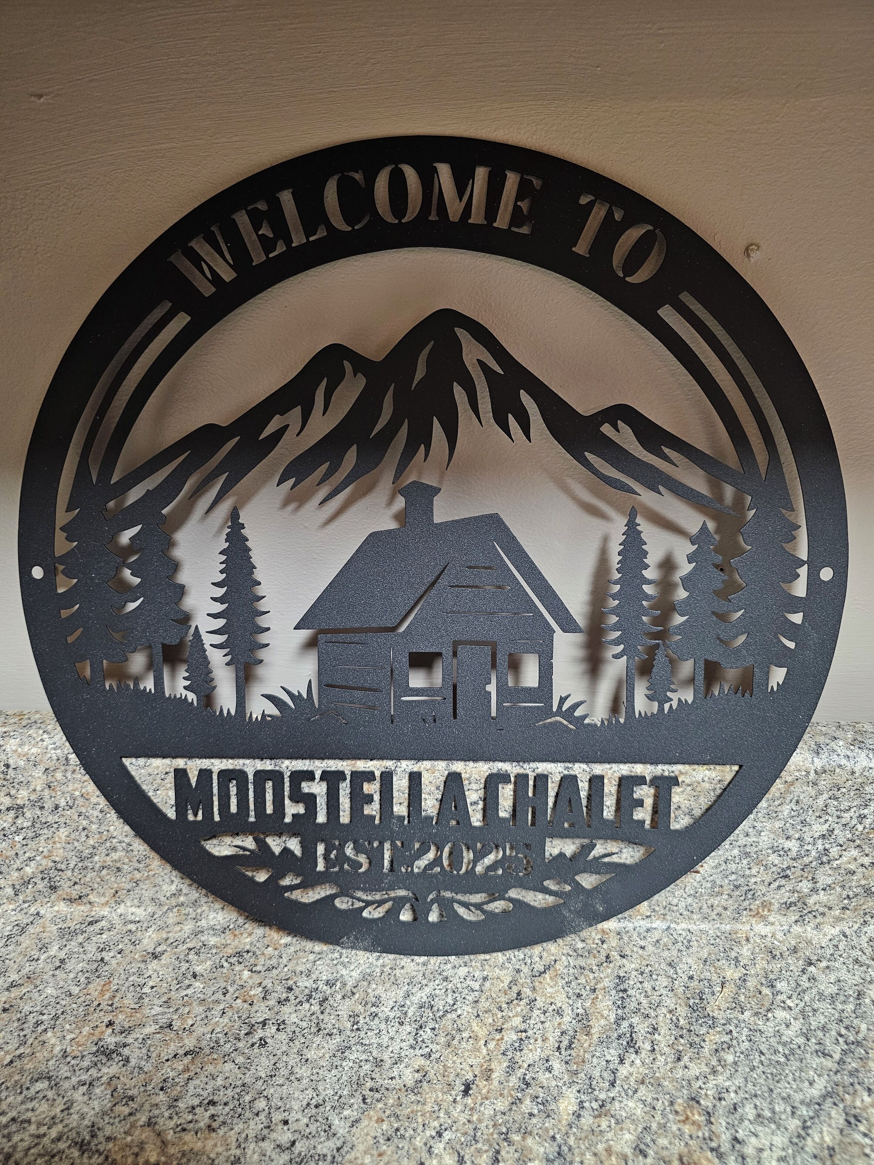 Welkomstbord voor Moostella Chalet, opgericht in 2025, met een huis omringd door bergen en bomen.