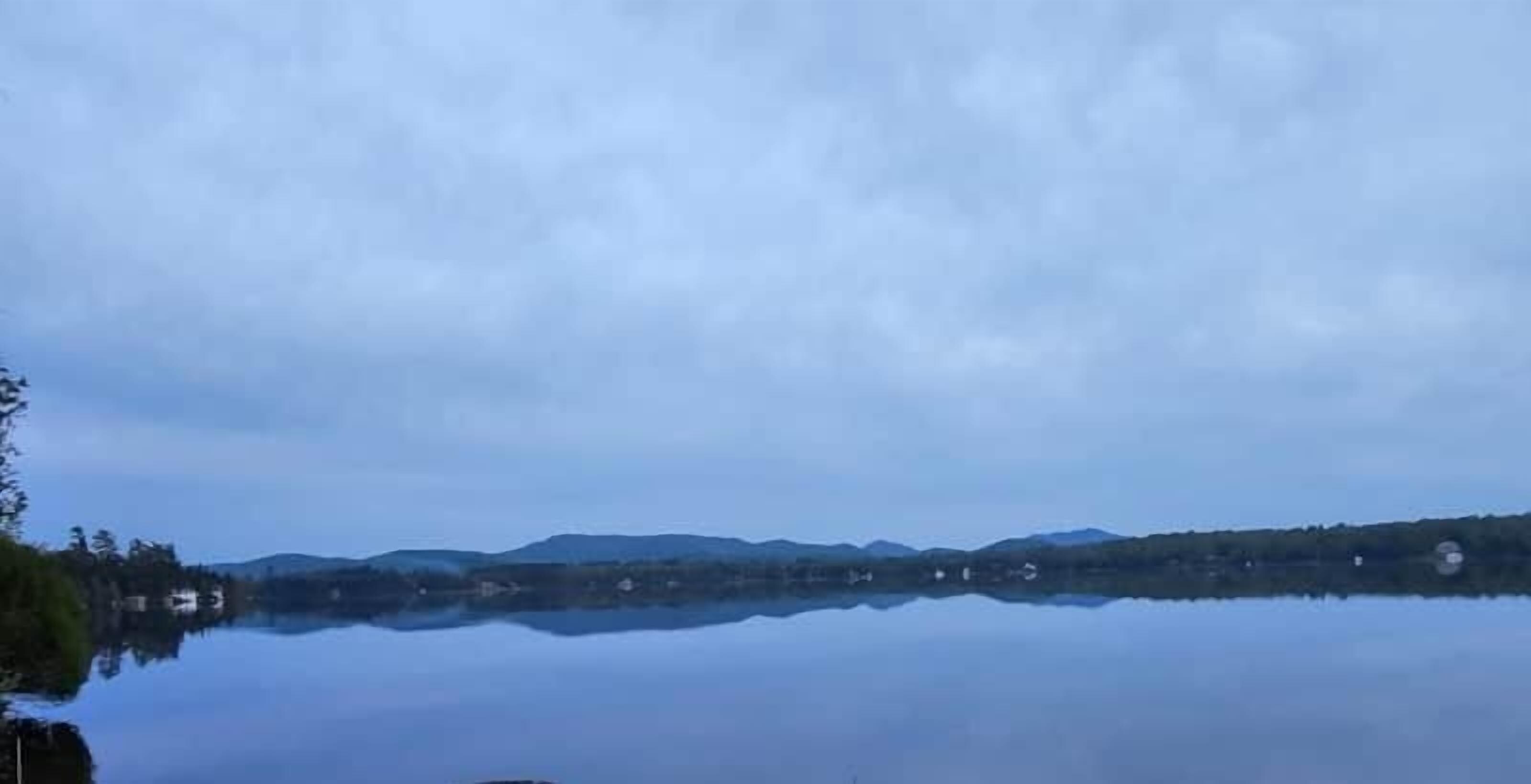 A calm lake with a clear reflection of the surrounding landscape, including distant hills and a few buildings.
