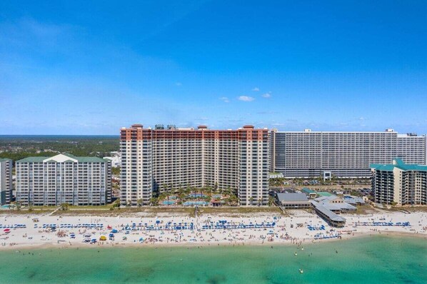 Un hotel frente a la playa con varios edificios altos, una playa arenosa con numerosas sombrillas y un cielo azul despejado.
