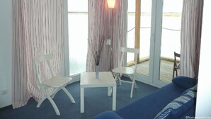 Une pièce avec un canapé bleu, deux chaises blanches, une petite table et une lampe. Des rideaux à rayures et une vue sur la mer par de grandes fenêtres agrémentent la pièce.