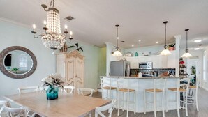 A dining area with a wooden table and white chairs, a chandelier, and a kitchen in the background.