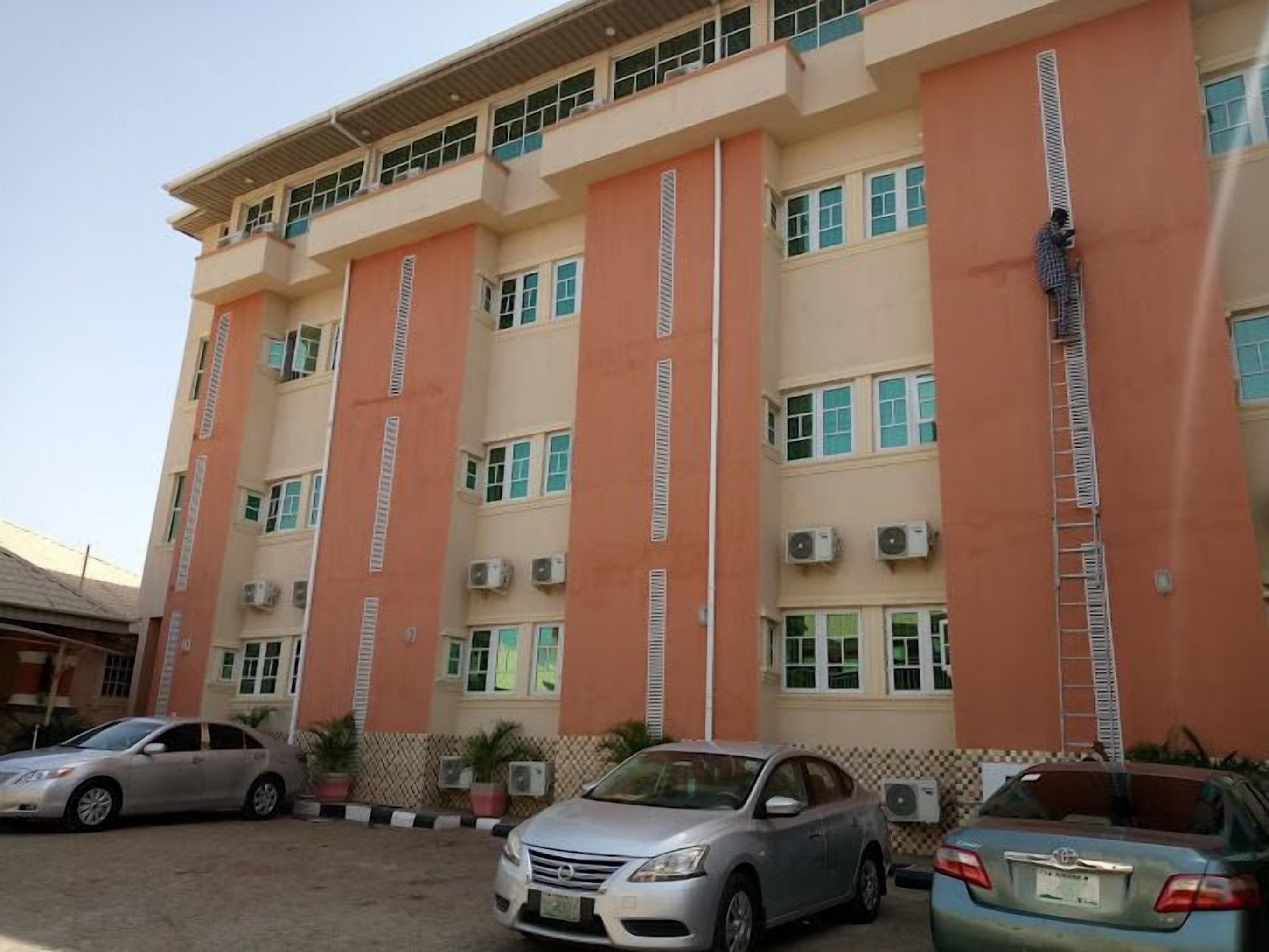 Un edificio de varios pisos con fachada color beige y naranja, unidades de aire acondicionado y un estacionamiento con coches.
