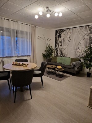 Ein modernes Wohnzimmer mit einem runden Esstisch, schwarzen Stühlen, einem grauen Sofa mit grünen Kissen und einem großen abstrakten Gemälde an der Wand.
