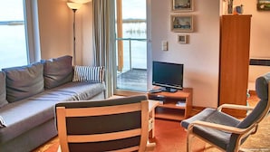 Ein gemütliches Zimmer mit einer Couch, einem Holzstuhl, einem Fernseher auf einem Schrank und Blick auf einen See durch das Fenster.