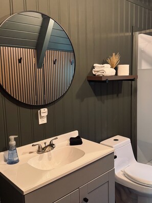 Une salle de bain avec un miroir rond, un lavabo avec un robinet, des toilettes et une étagère avec des serviettes et une plante.