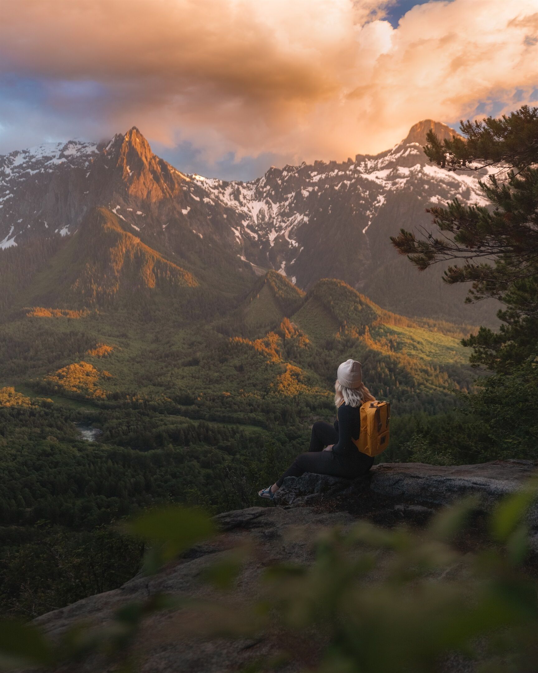Eine Person sitzt auf einem Felsen mit Rucksack und blickt auf eine gebirgige Landschaft mit schneebedeckten Gipfeln und einem bewaldeten Tal.