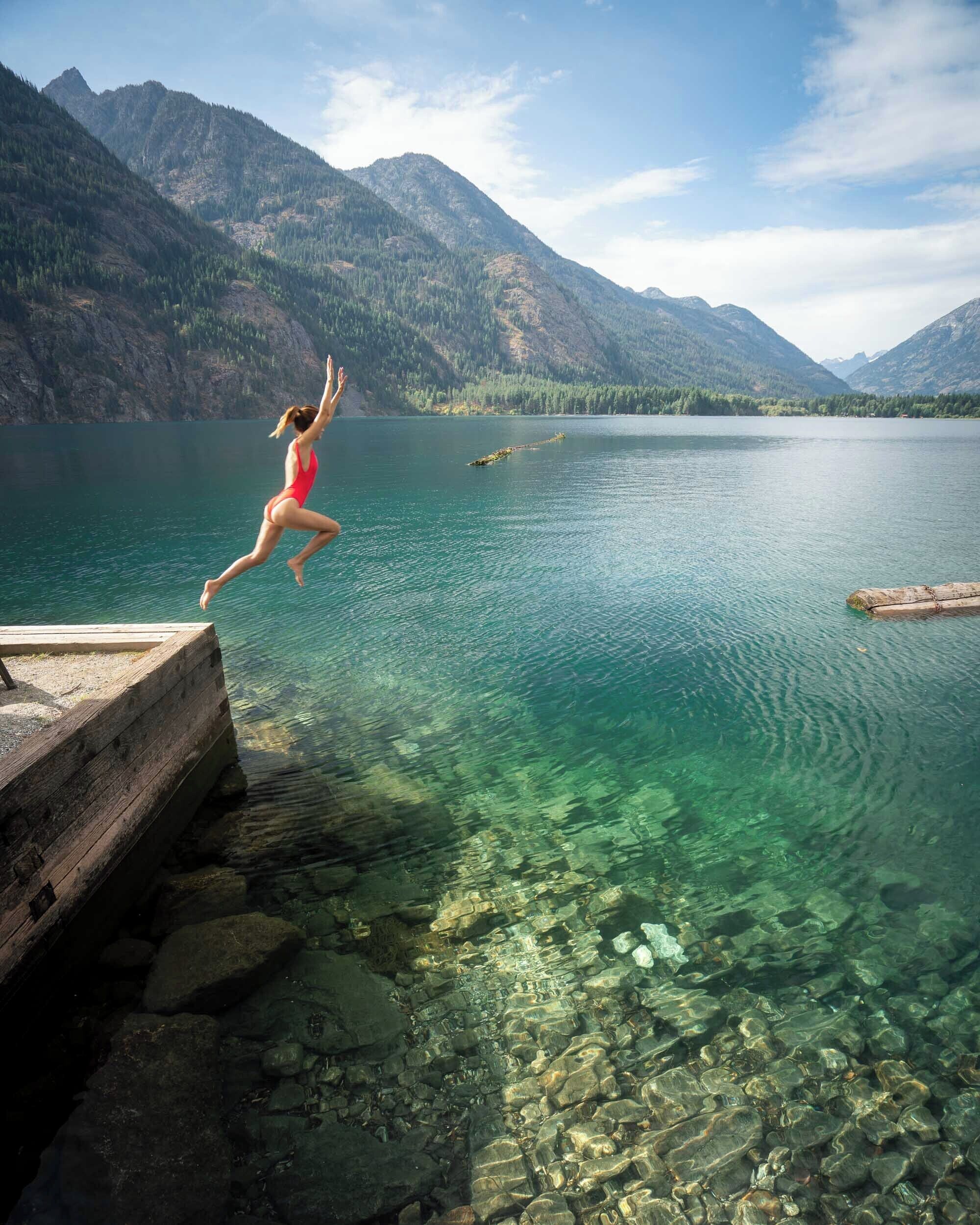 A woman jumping off a wooden dock into a clear lake surrounded by mountains.