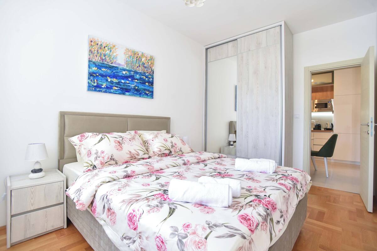 A bedroom with a bed, a nightstand, a painting, and a view into the kitchen.