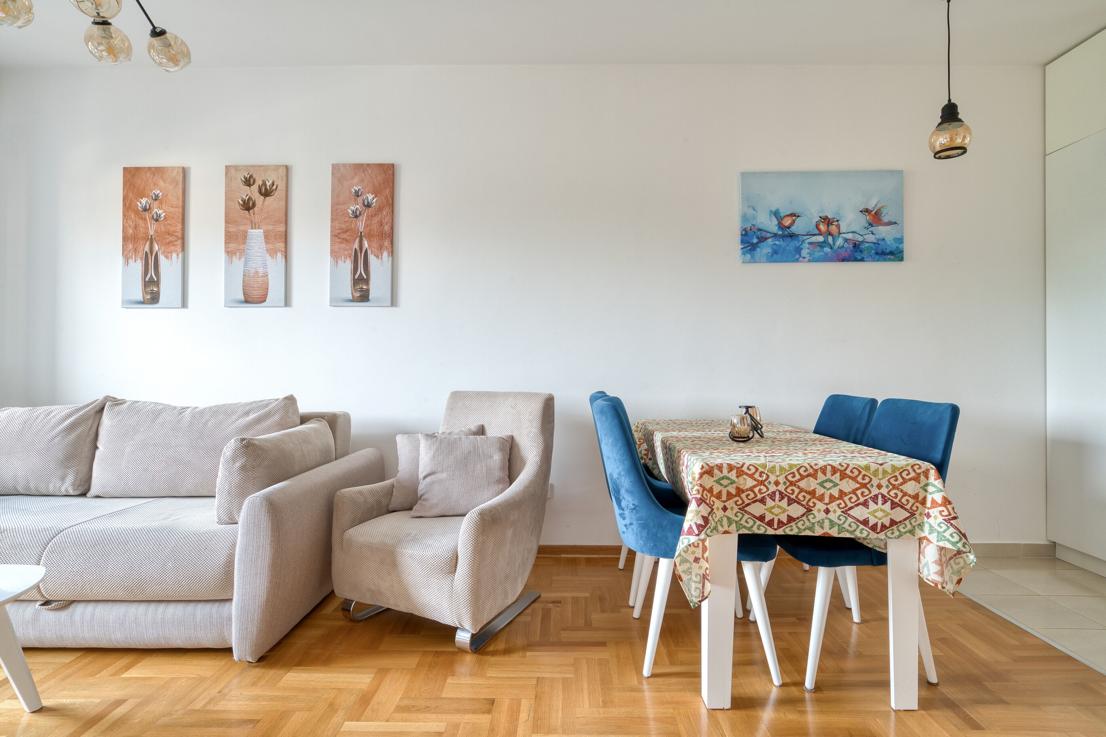 A modern living room with a sofa, armchair, and dining table with chairs. There are three paintings on the wall.