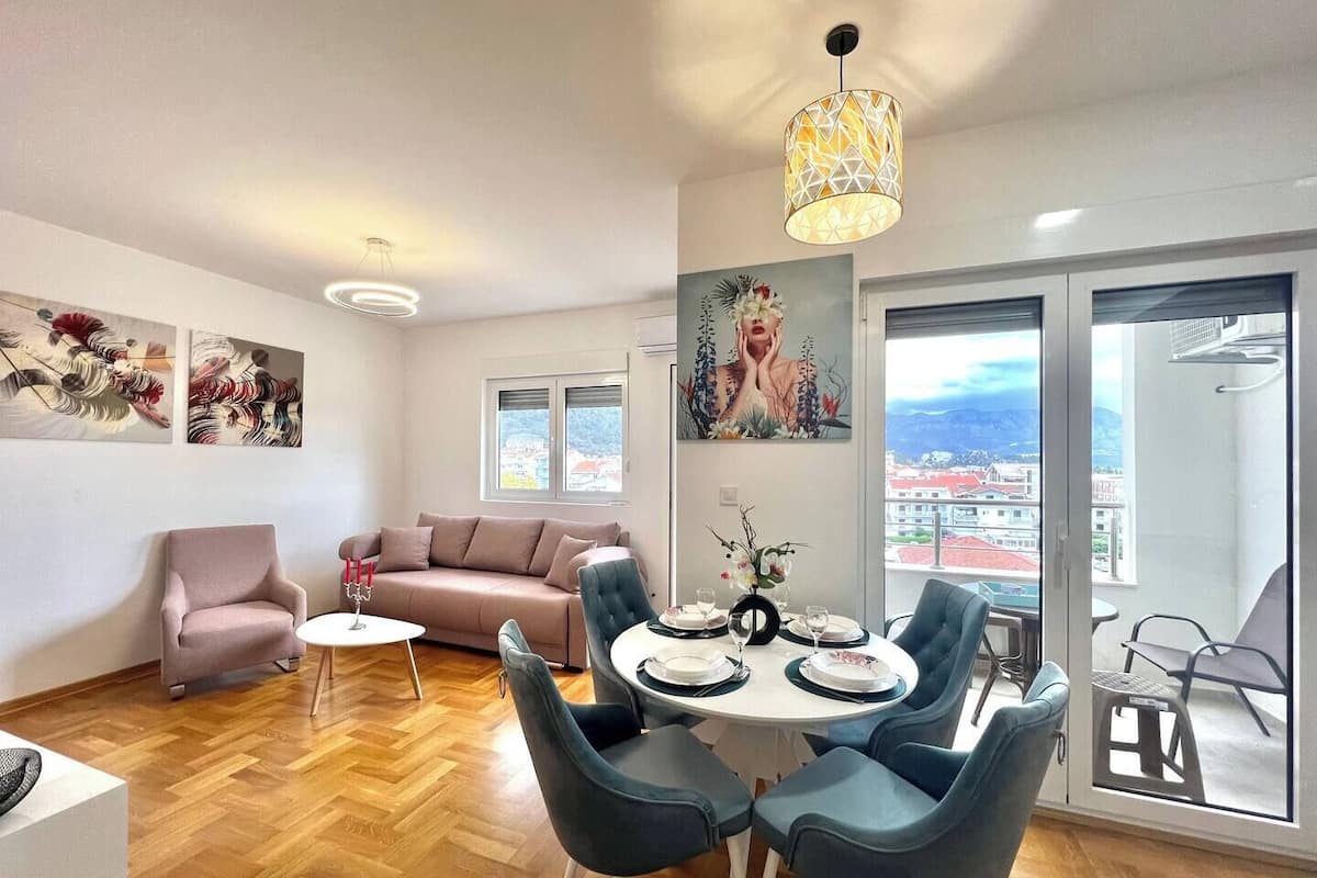 A modern living room with a dining area, a sofa, a coffee table, and a chandelier. There are two windows with a view of buildings and mountains.