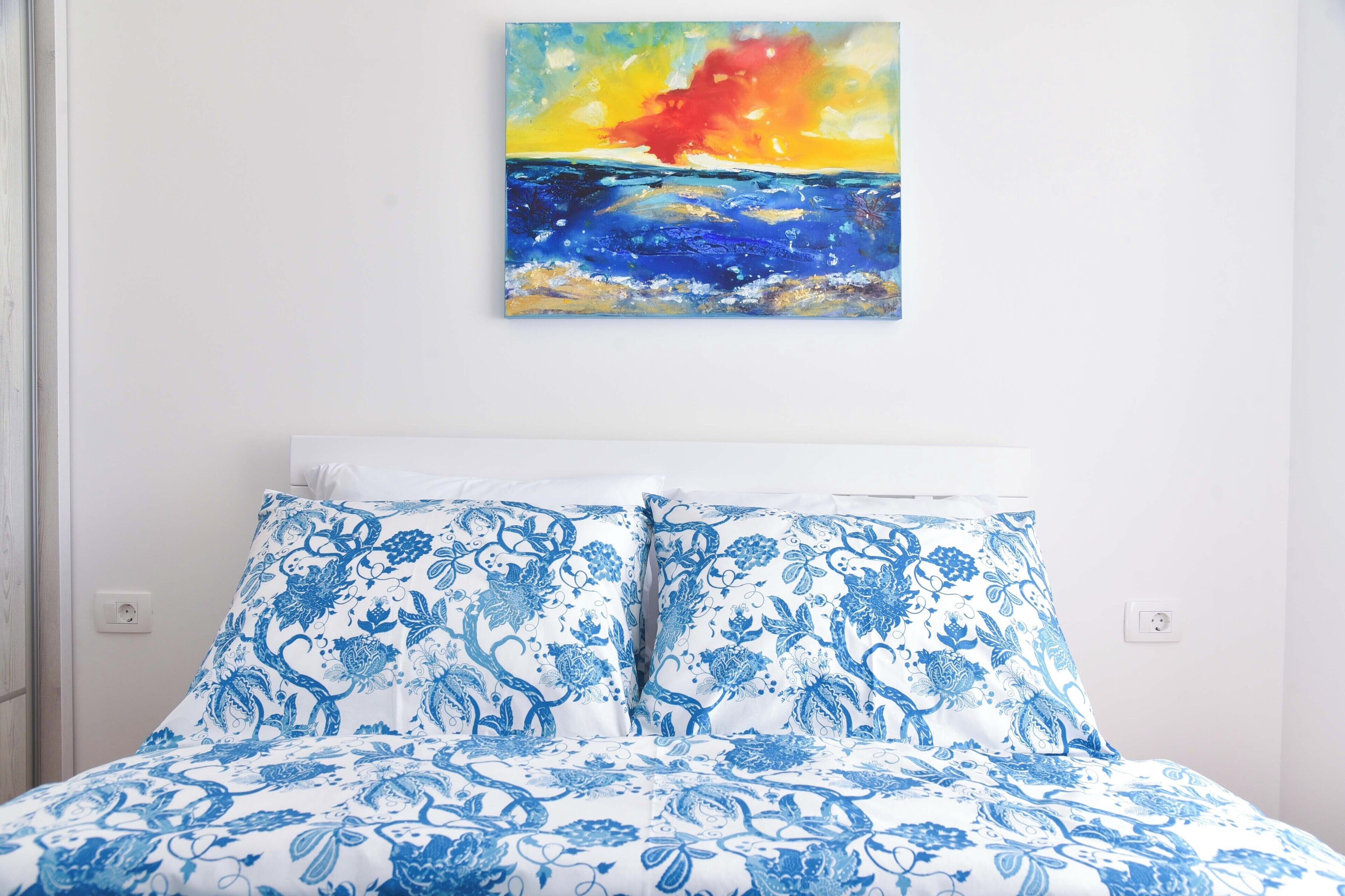 A bed with blue floral bedding, a white headboard, and a painting of a sunset over the ocean on the wall.