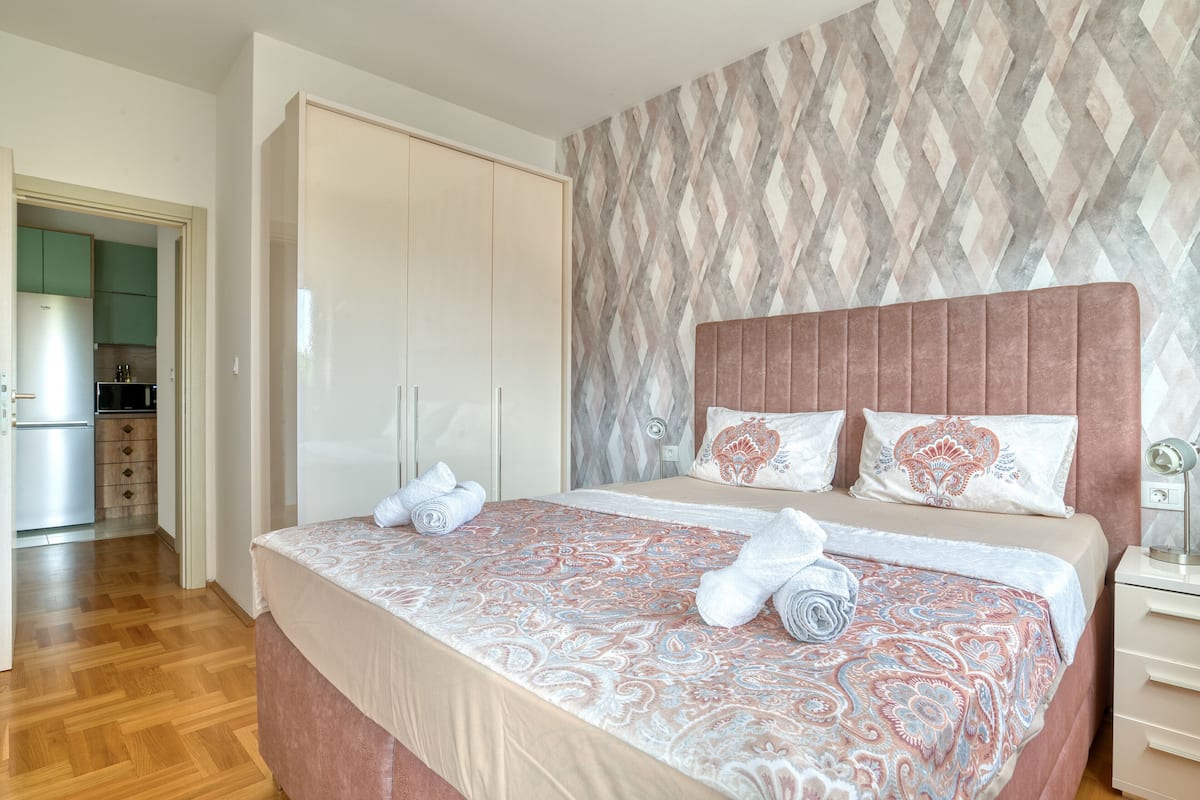 A bedroom with a large bed, a wardrobe, and a view into the kitchen.