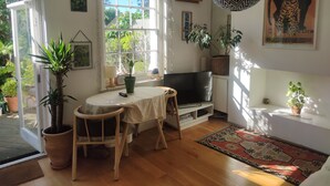 A cozy living room with a wooden floor, a round table, a TV, and various plants.