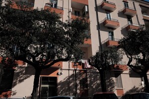 A multi-story building with balconies, trees in front, and parked cars.