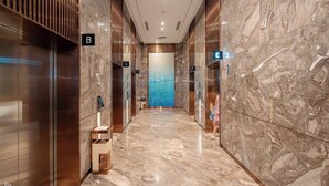 Corredor moderno de hotel com pisos de mármore, portas de elevador identificadas com as letras B, C, D, E e F, e uma grande obra de arte azul na parede.
