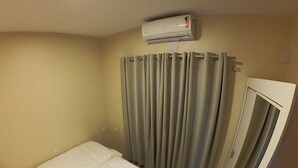 Une petite chambre avec un lit simple, un lit blanc, des murs de couleur beige, un luminaire au plafond, une fenêtre avec des rideaux et un climatiseur fixé au mur.