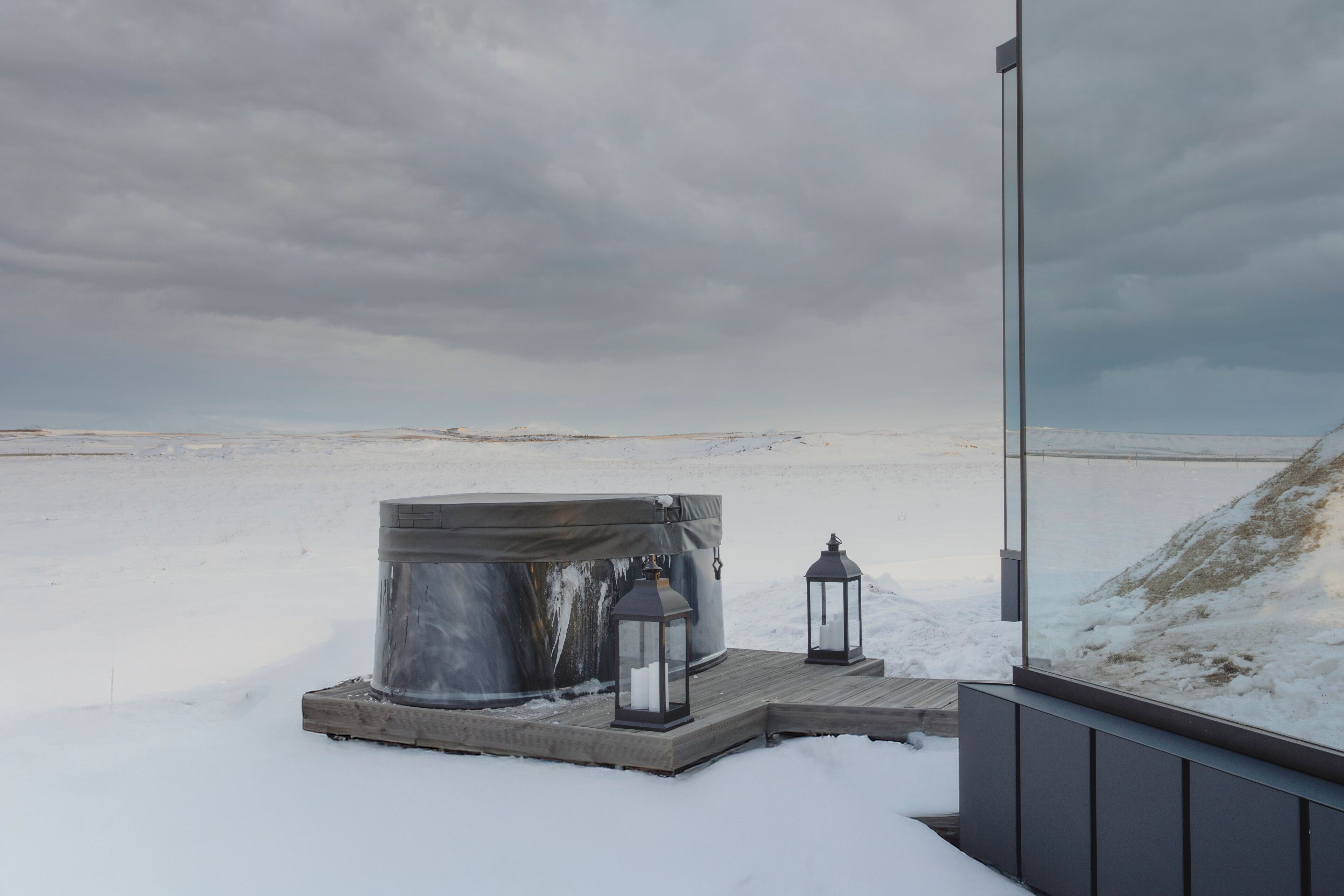 A hot tub on a wooden platform with two lanterns, set against a snowy landscape and cloudy sky.
