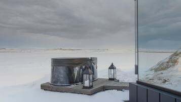 A hot tub on a wooden platform with two lanterns, set against a snowy landscape and cloudy sky.