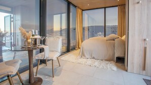 Une chambre moderne avec une grande fenêtre, un lit, une chaise et une table ornée d’un vase et de bougies.