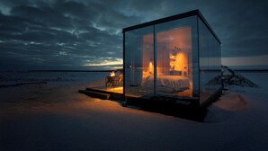 Une cabane aux parois de verre, comprenant un lit, une petite table et une chaise, située dans un paysage enneigé et offrant une vue sur l’horizon.