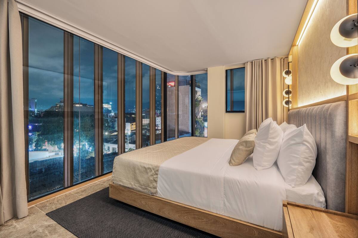 A modern hotel room with a large bed, a city view through floor-to-ceiling windows, and a wooden headboard with ambient lighting.