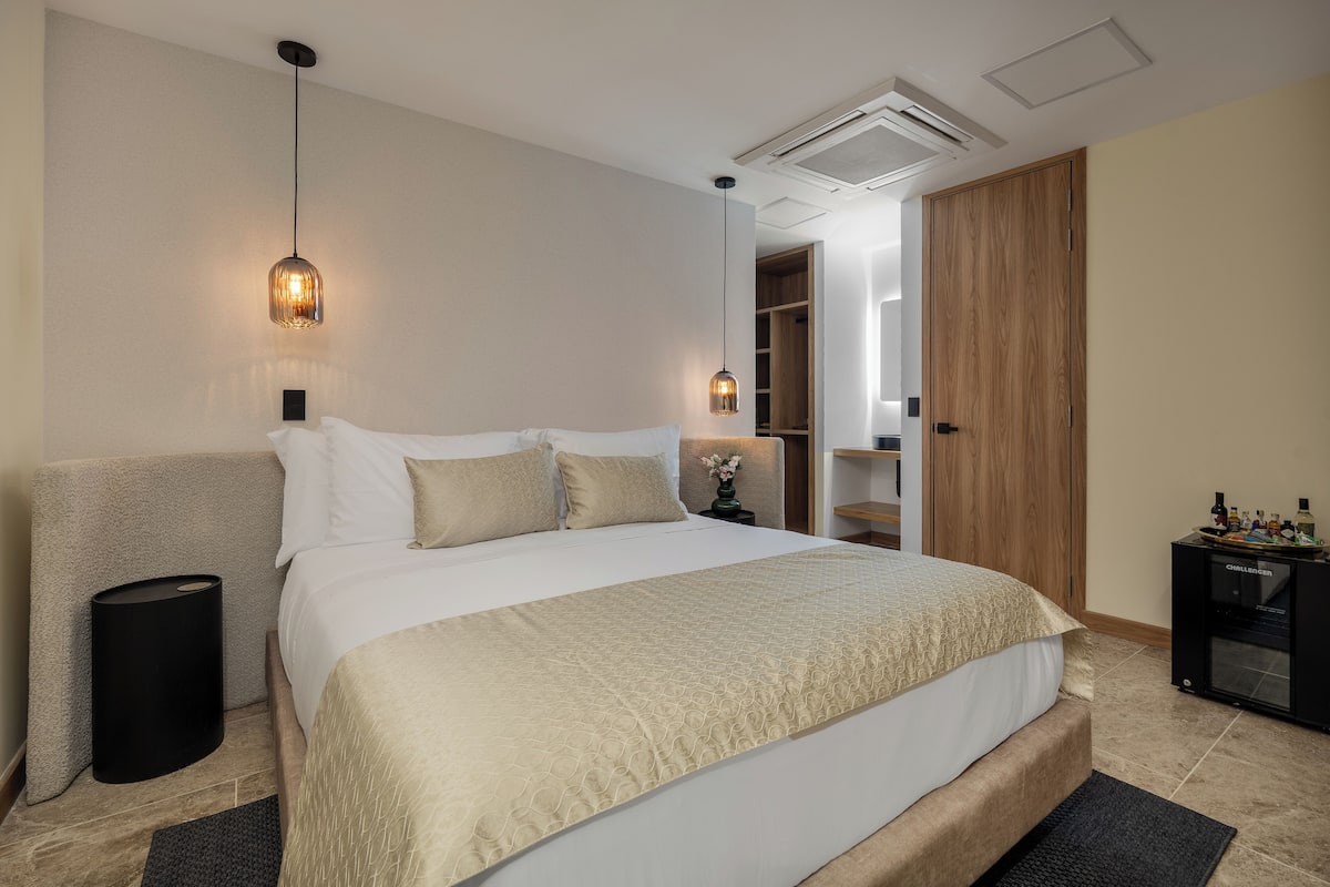 A modern hotel room with a large bed, two pendant lights, a built-in wardrobe, and a minibar.