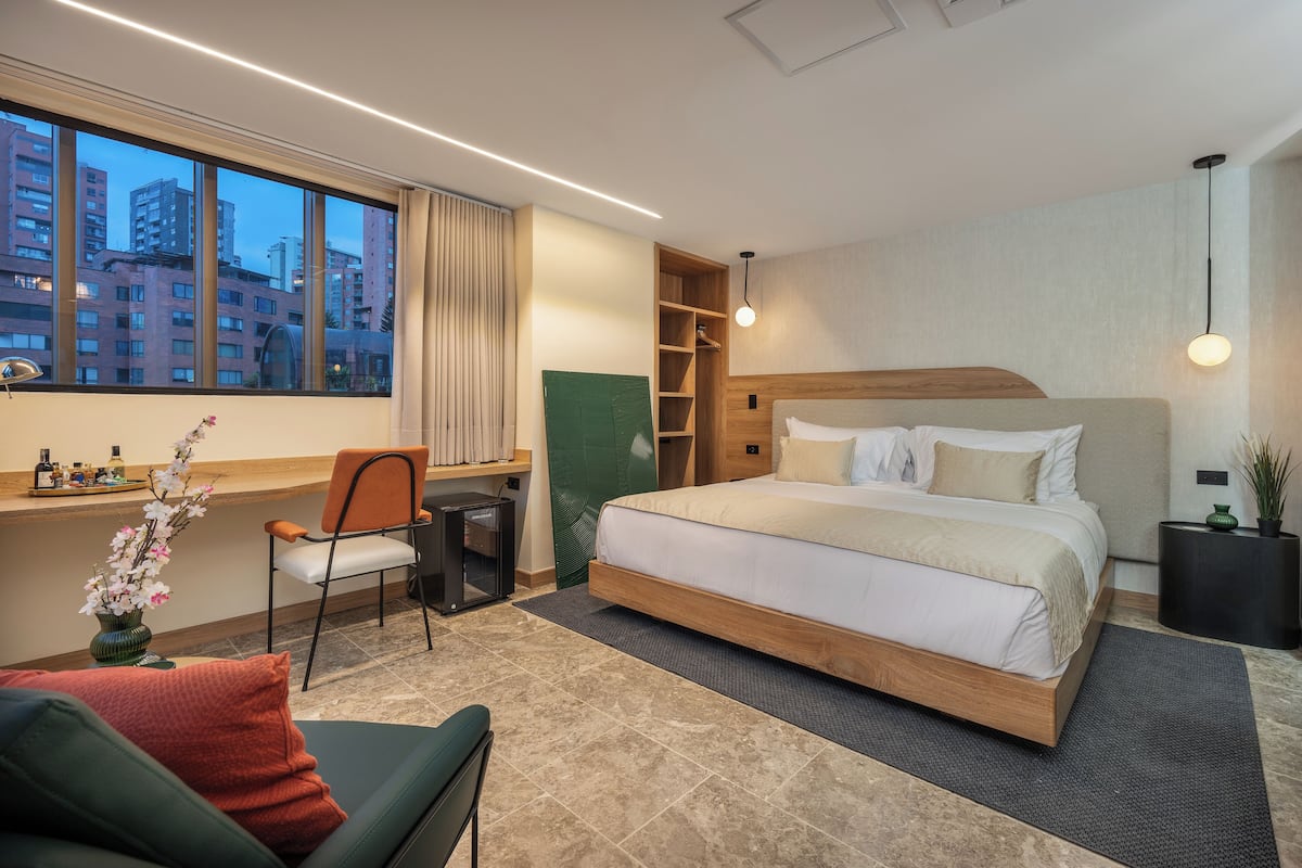 A modern hotel room with a large bed, a desk with a chair, a small table, and a view of tall buildings through the window.