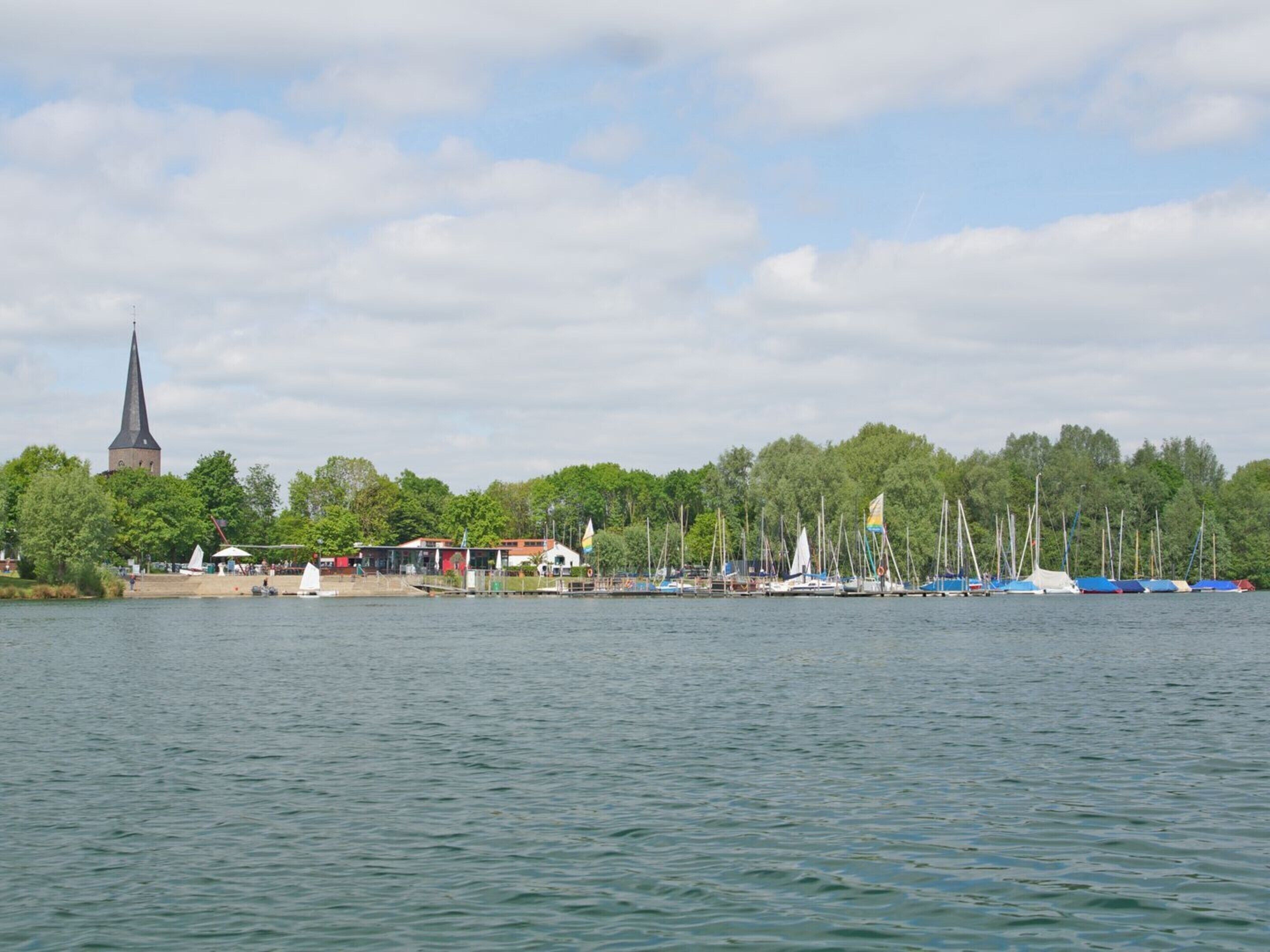 A waterfront view with a church spire, sailboats, and a forested area.