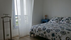 A bedroom with a bed, a nightstand, a lamp, a window with sheer curtains, and a wooden hanger rack. - Apartment rental, sea view, Port-Louis (56), 3-star CLEVACANCES label (Port-Louis)