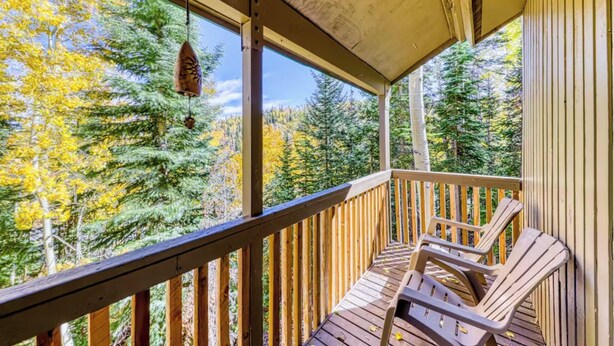 A wooden deck with two Adirondack chairs, a hanging bird feeder, and a view of a forest with autumn foliage.