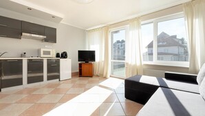 A modern kitchen with a microwave, stove, and flat-screen TV. There is a large window with curtains, a sofa, and a view of buildings outside. - Comfortable holiday studio, breakfast included (Rewal)