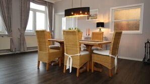 Un coin repas avec une table ronde, quatre chaises en osier dotées de coussins blancs, un luminaire suspendu et une fenêtre avec des rideaux.
