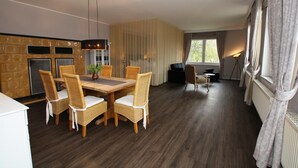 Un salon moderne avec un coin repas, comprenant une table à manger en bois, des chaises en osier, un canapé et une grande fenêtre avec des rideaux.