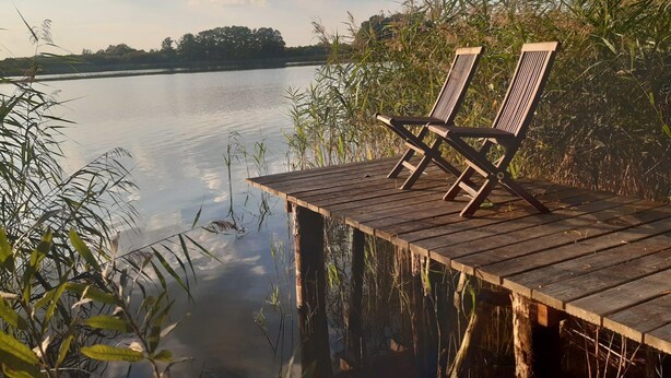 Une jetée en bois avec deux chaises, donnant sur un lac paisible, le tout entouré de végétation.