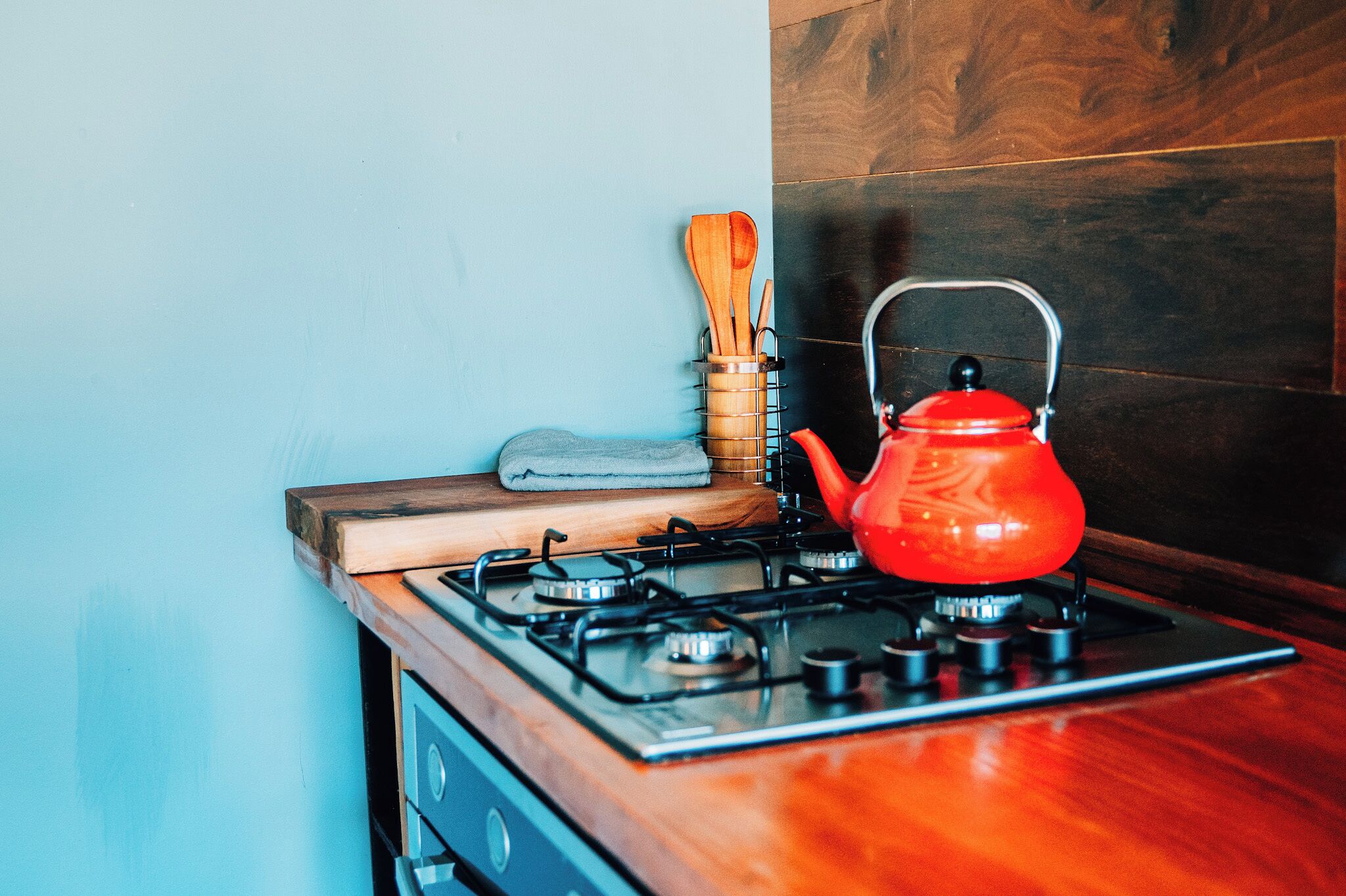 Una cocina con un hervidor rojo sobre la estufa, una tabla de cortar de madera y un juego de utensilios de cocina de madera.