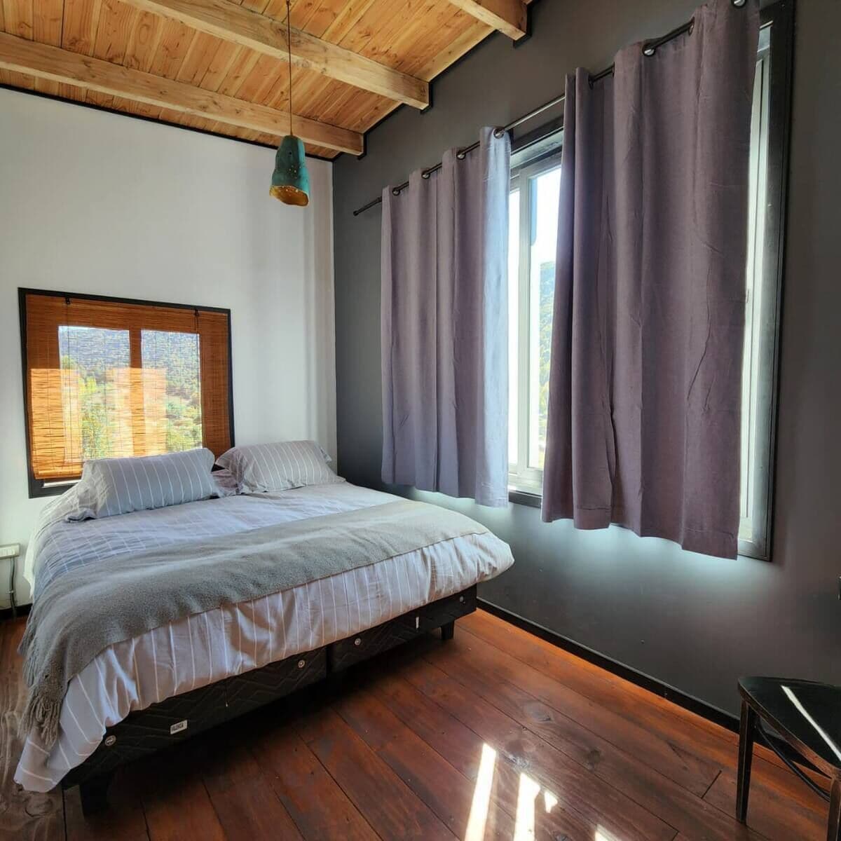 A bedroom with a wooden ceiling, a bed with a grey and white striped duvet, a wooden floor, and a window with purple curtains.