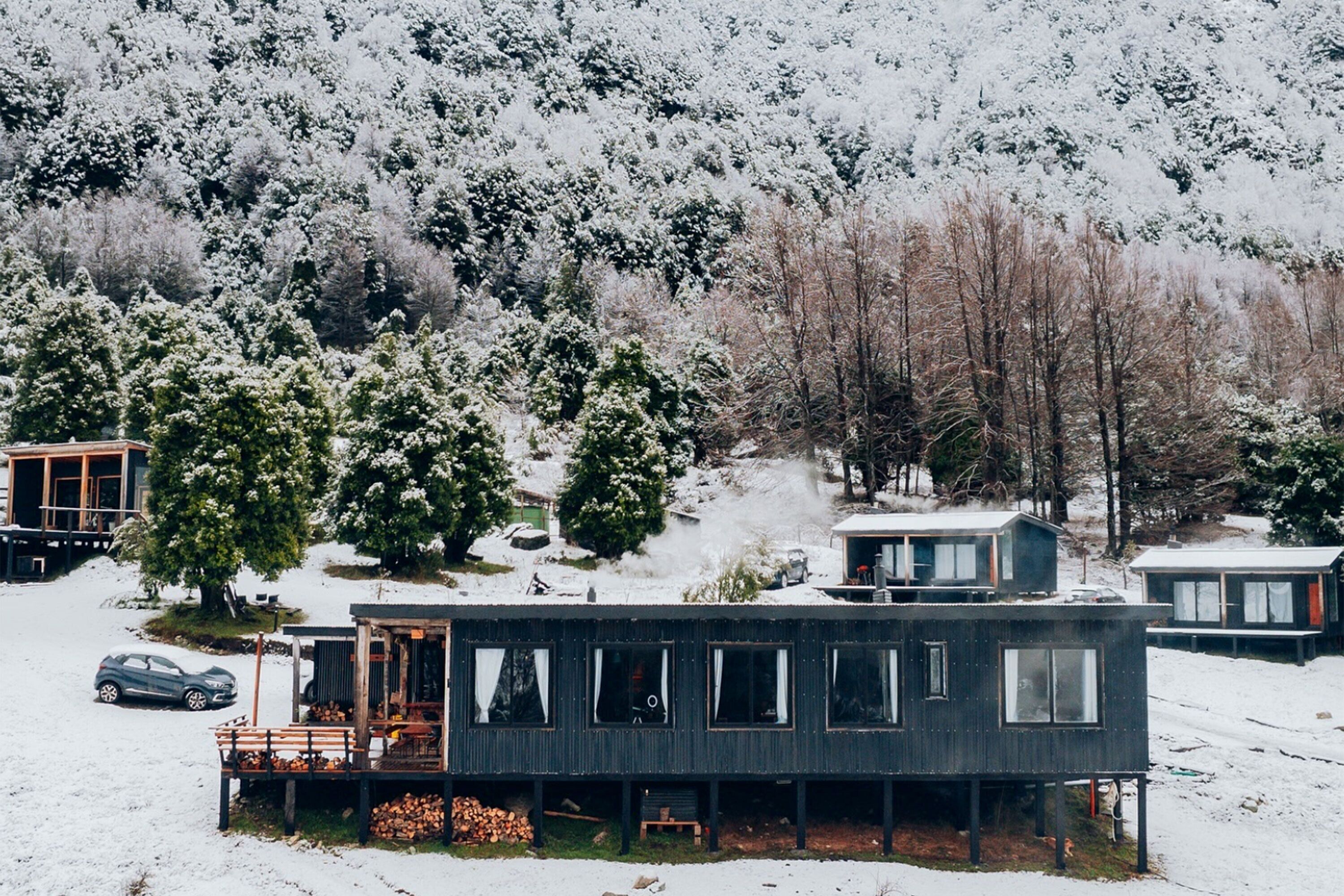 A modern cabin-style building with multiple rooms, situated on a snow-covered hillside with trees in the background. - K10 Lodge (Curacautin)