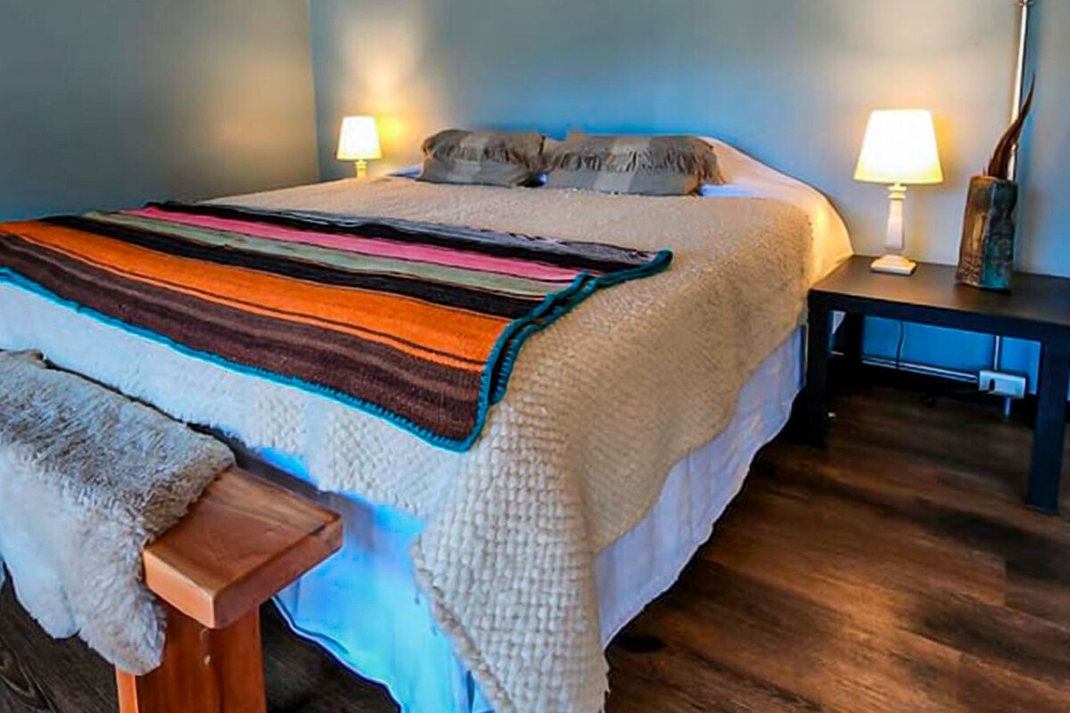 Un lit bien fait, recouvert d’une couverture à rayures, avec deux lampes de chevet et un banc en bois agrémenté d’un plaid en fourrure.