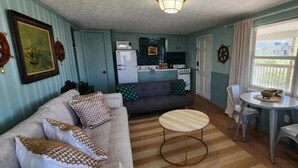 Un espace de vie compact comprenant un canapé, une table basse ronde, une table à manger avec des chaises, un coin cuisine et une fenêtre avec des stores.