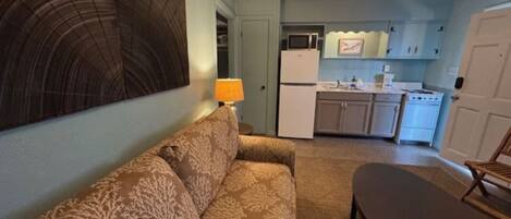 A compact living space with a sofa, a small table, a lamp, and a kitchenette with a refrigerator, microwave, and stove.