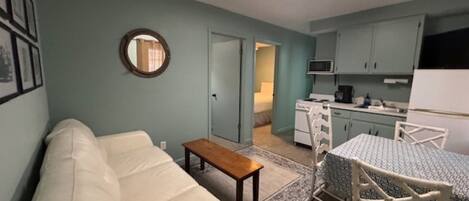 Habitación pequeña con cama, una mesita, kitchenette y baño a la vista a través de una puerta abierta.